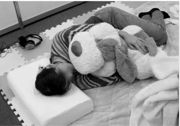 図 2.1  抱き枕型システム ZZZoo Pillows を使用している様子 2.2.  抱き枕型システムで扱う生理情報  疑似的な添い寝をしているような感覚を与える抱き枕型のシステムを開発するにあたり， 呼吸時の胸郭の動きや体温などの生理情報に着目した．入眠を目的とする状況に対し，入 眠目的で瞼を閉じることにより視覚情報は遮断されることから，表情や視線や身振りなど， 視覚によった情報の提示は，不向きであると考えられる．特に呼吸は，渡辺ら [33 ， 34] の実 験において，呼吸の引き込み現象が円滑なコ